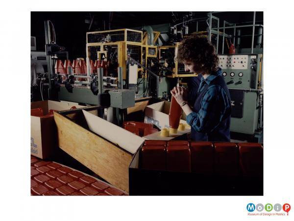 Scanned image showing a female worker adding tops to bottles on a production line.