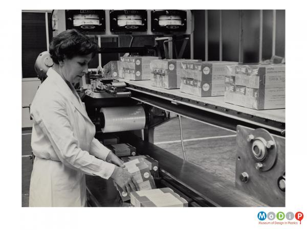 Scanned image showing a female worker preparing boxes to be shrink wrapped on a production line.