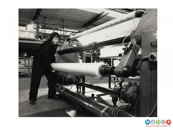 Scanned image showing a male worker handling a large roll of material, either removing or adding it to a machine.
