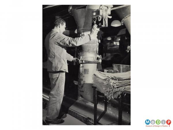 Scanned image showing a lined hessian sack being filled by a machine controlled by a male worker.