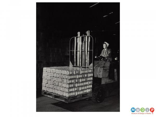 Scanned image showing a female worker driving a forklift truck loaded with a pallet full of shrink wrapped jars.