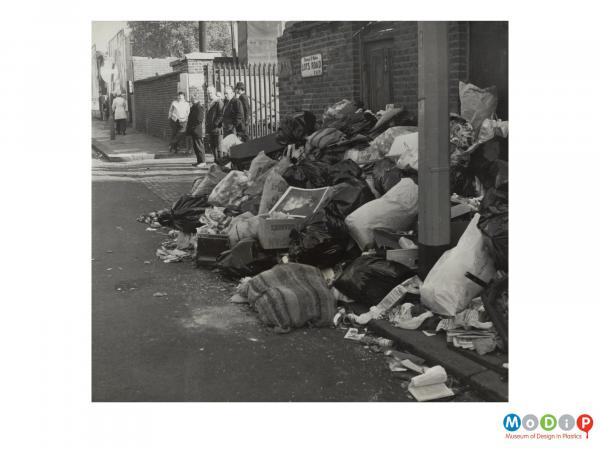 Scanned image showing a street scene depicting piles of filled rubbish bags.