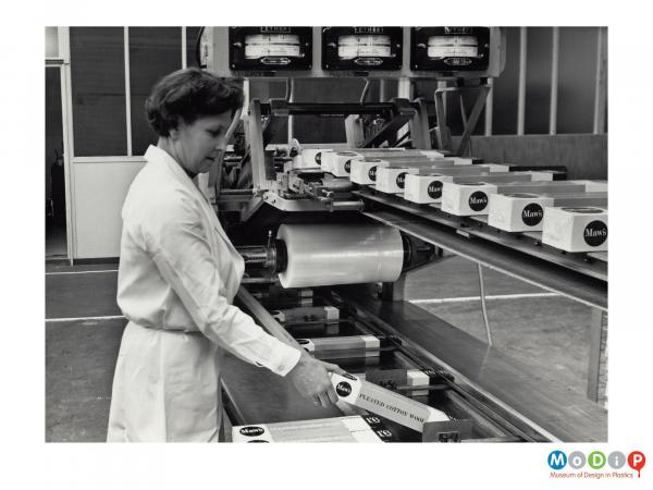 Scanned image showing a woman putting boxes into a wrapping machine.