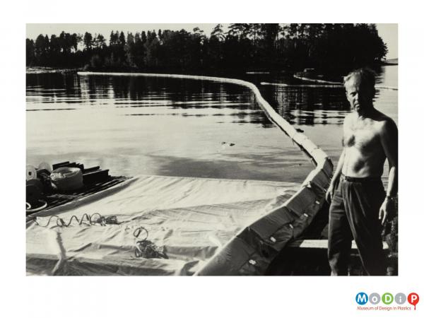 Scanned image showing a man standing near a boom flaoting in the water behind him.