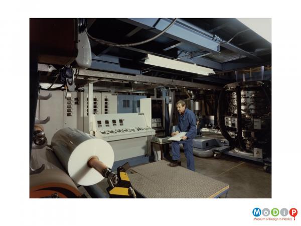 Scanned image showing a man in blue overalls taking notes by an extruding machine.