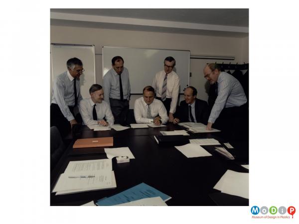 Scanned image showing a group of male employees gathered by a meeting table.