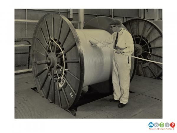 Scanned image showing a male worker feeding coiling a cable onto a large drum.