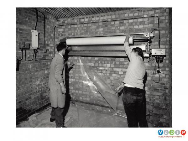 Scanned image showing 2 men cutting a sheet of plastics material from a roll mounted on a wall.