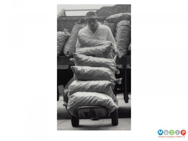 Scanned image showing a man pushing a pile of sacks on a sack truck.
