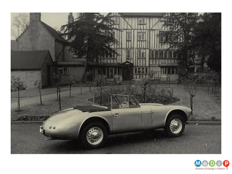 An MG sports car outside a large Tudor style house.  The car body is in an unfinished state.